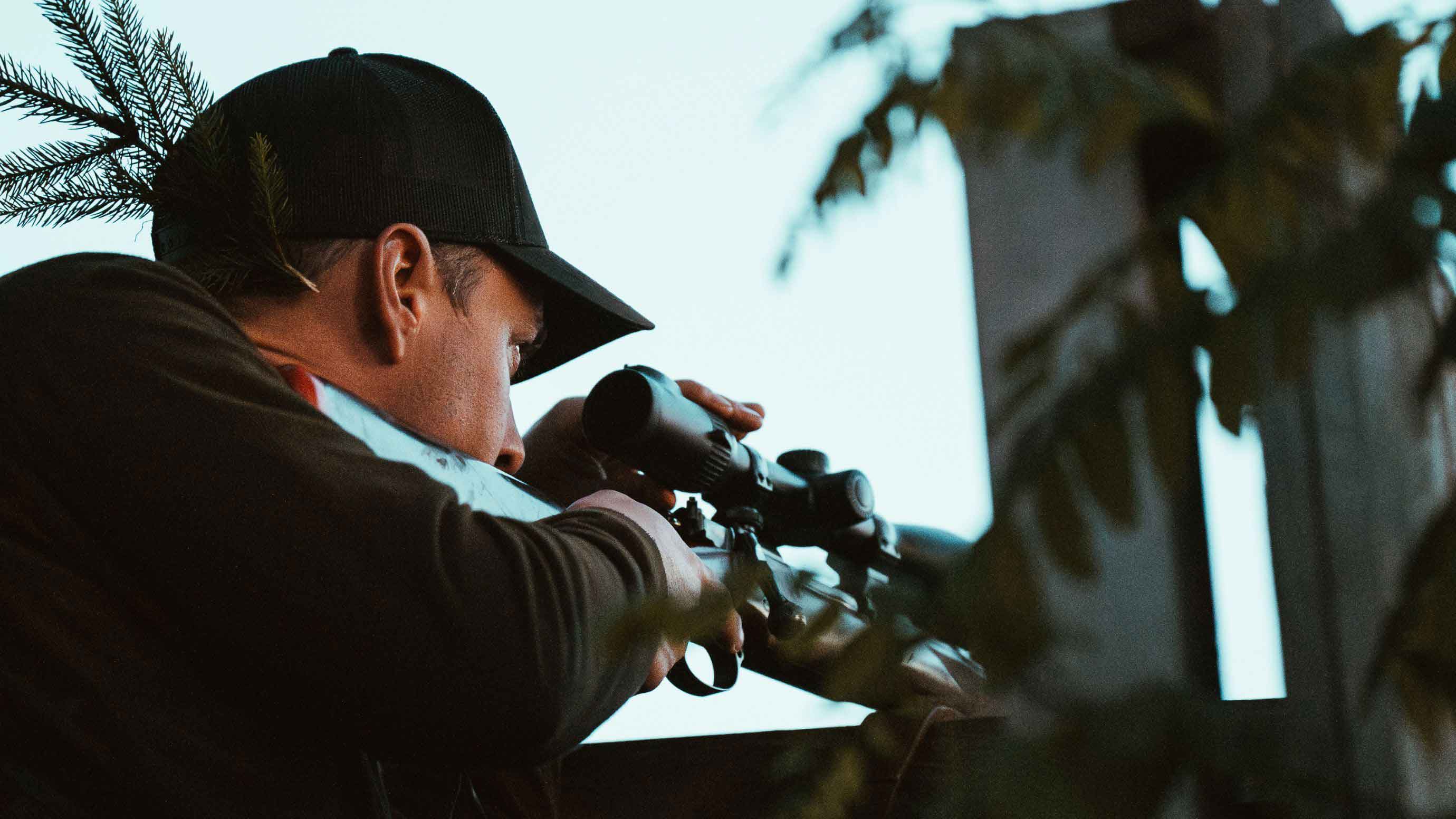 Eine Person von hinten, die ein Gewehr mit Zielfernrohr hält und auf ein unsichtbares Ziel zielt.