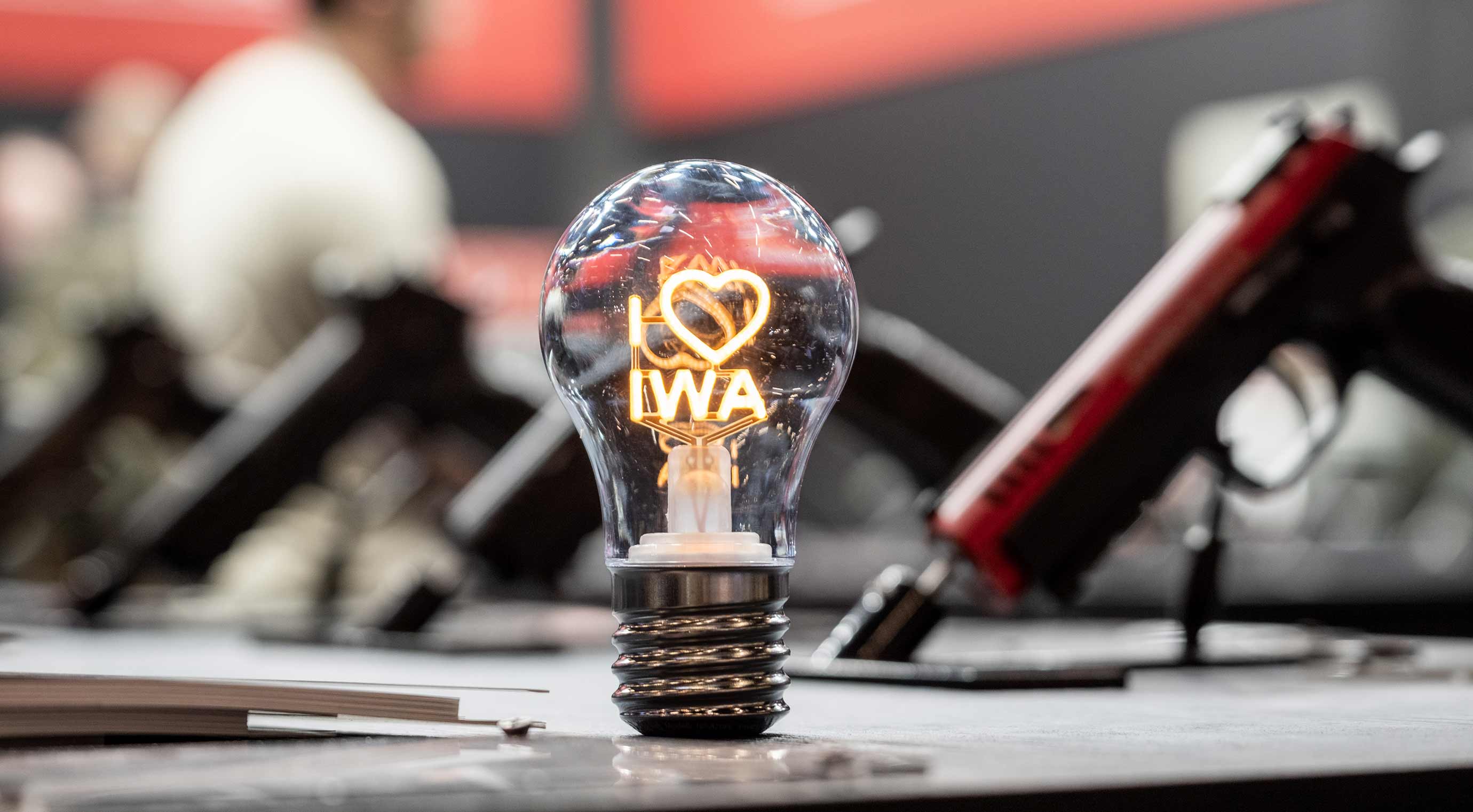 Glühbirne mit dem leuchtenden Schriftzug ‚I love IWA‘ steht auf einem Messetresen. Im Hintergrund sind unscharf ausgestellte Waffen zu sehen.