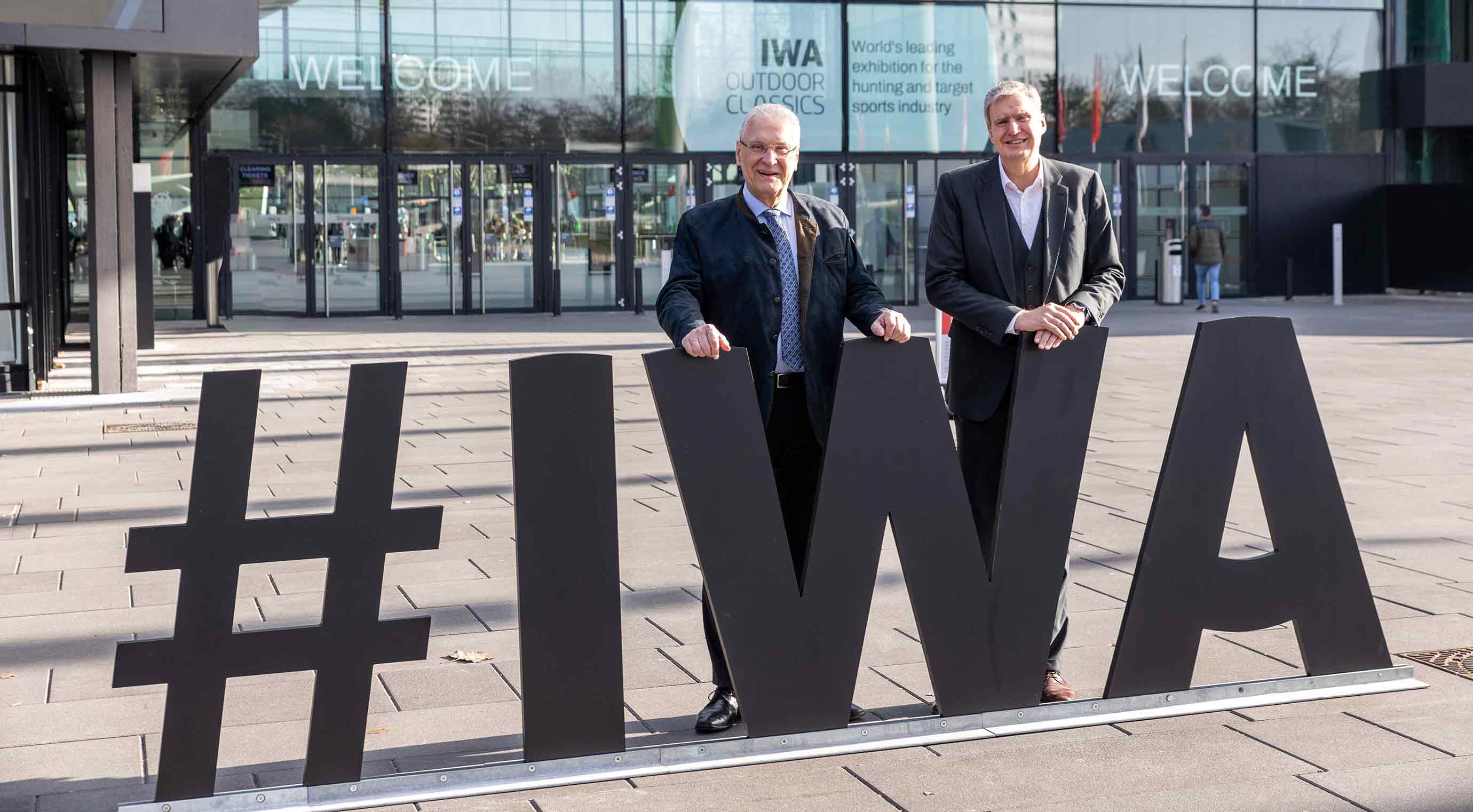 Joachim Herrmann und Peter Ottmann stehen vor dem Eingang der IWA neben einem großen, schwarzen #IWA‑Schriftzug. Im Hintergrund ist der Eingangsbereich mit Welcome‑Beschriftung und Messegrafiken zu sehen.