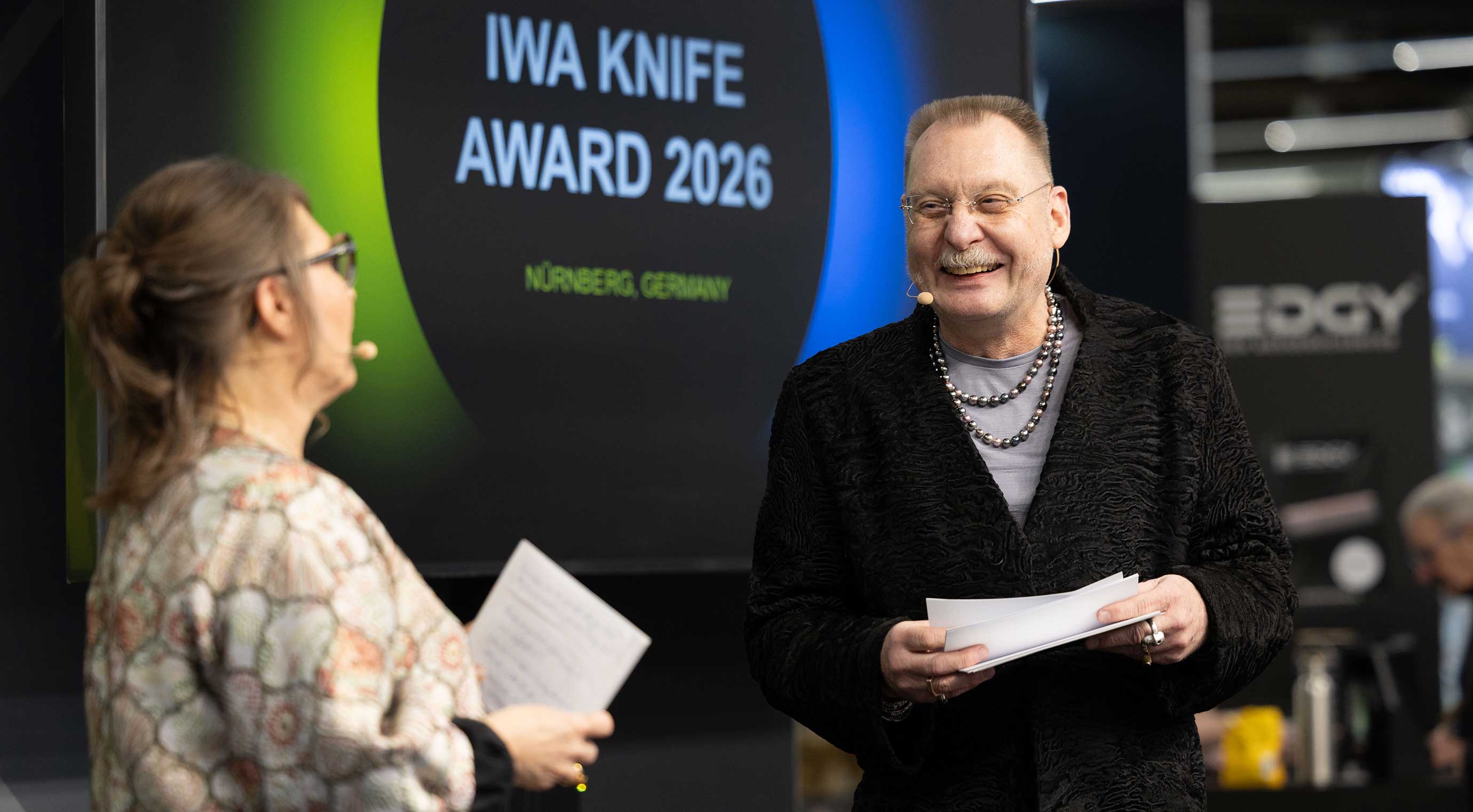 Zwei Personen stehen auf einer Bühne. Im Hintergrund ist eine große Leinwand mit dem Schriftzug ‚IWA Knife Award 2026 – Nürnberg, Germany‘ zu sehen.